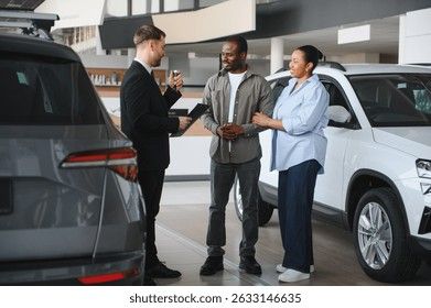 A car salesman in a suit hands over keys to a smiling couple in a showroom.